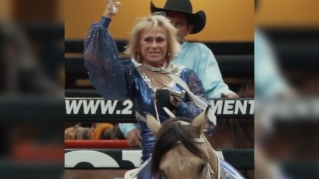 A 72-year-old woman smiles and raises a hand in the air as she rides a horse