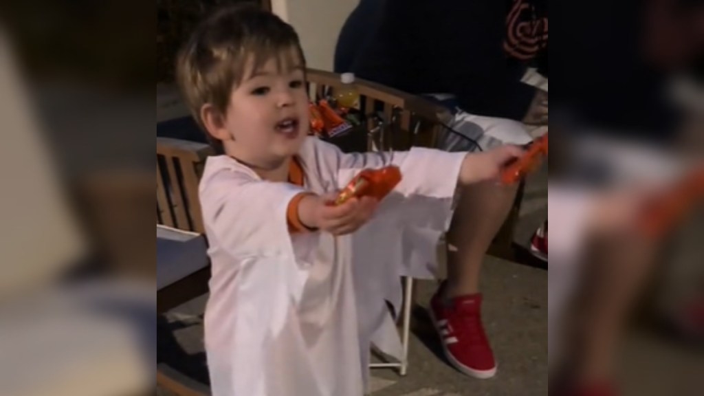 Close up of a little boy standing outside at night, an adult sitting next to him. The little boy is stressed in white, like a ghost. He talks as he holds out candy in each hand.