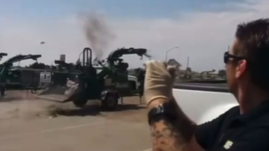 A man holds up his phone toward a wood chipper that's in the distance. The wood chipper seems to be in use