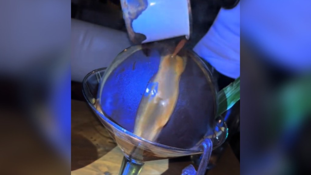 Close up of a dessert. A late chocolate ball sits in a large drinking glass. Warm liquid is being poured onto the chocolate.
