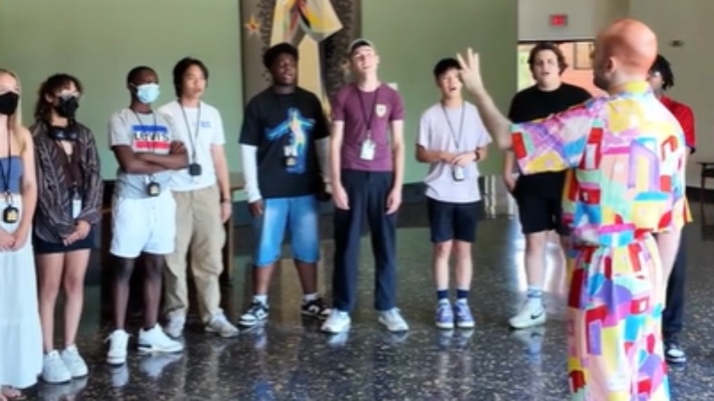 A choir of young people line up next to one another and sing. Their choir director stands in front of them, holding up a hand with three fingers raised