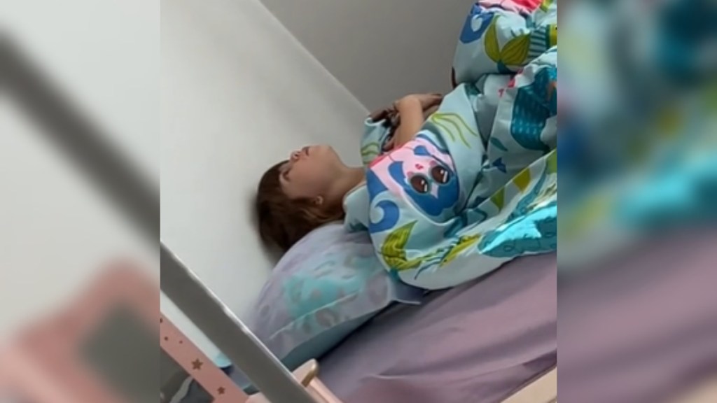 View from a distance of a little girl laying in bed on her back. She looks up at the ceiling, mouth open because she's singing