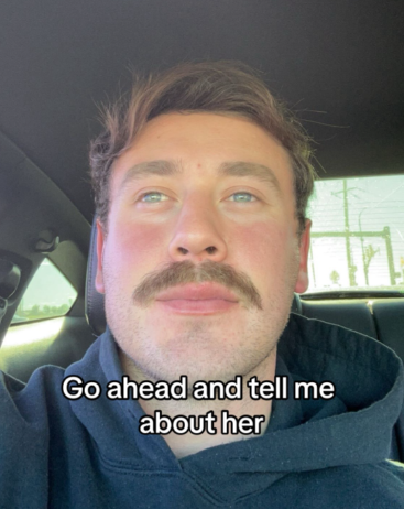 A man takes a selfie in the car. He looks away from the camera and at something in the distance.
Text on the image reads: Go ahead and tell me about her