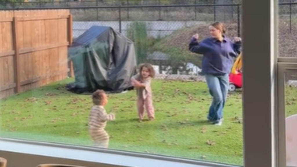 A view of a backyard, from a distance, and through a window. Outside, a woman dances next to her two little girls, who are also dancing