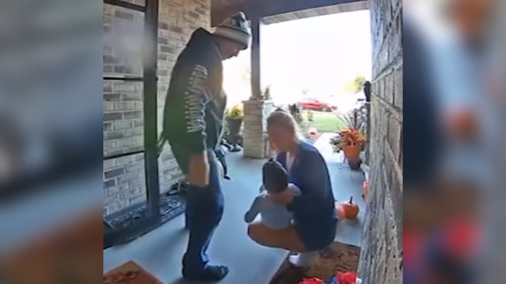 View of a front porch from the doorbell cam. A woman squats with her child in her arms. She looks down at him. A man stands next to them, door still open as he joins them.