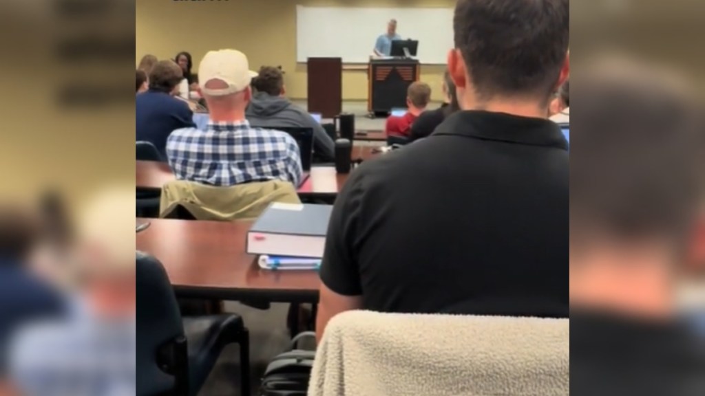 View from a student sitting in class. There are rows of tables and students and a professor standing at the front of class in front of a podium/desk that has a computer on it