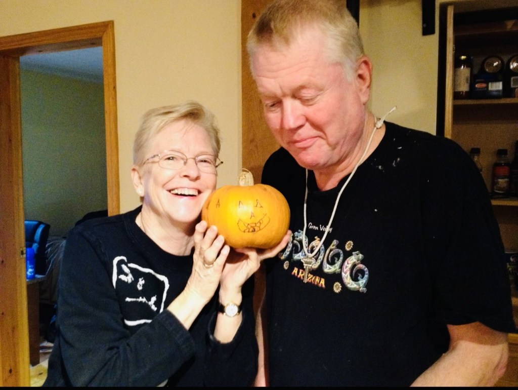 Kathleen holds up a miniature pumpkin while smiling next to Ed