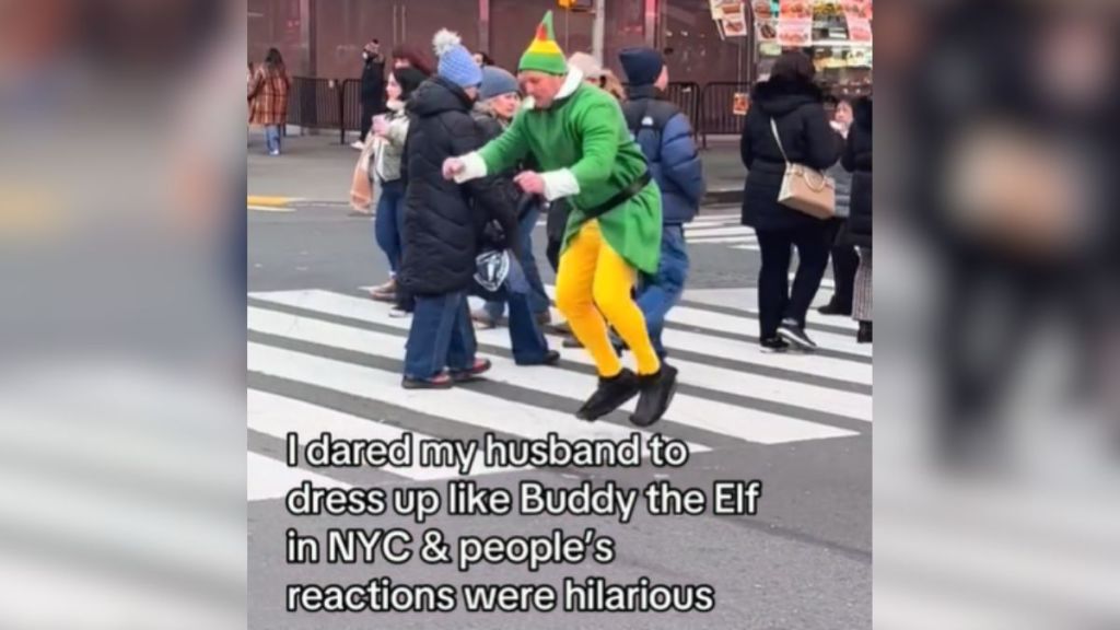A man hopping across a busy crosswalk dressed as Buddy the Elf.