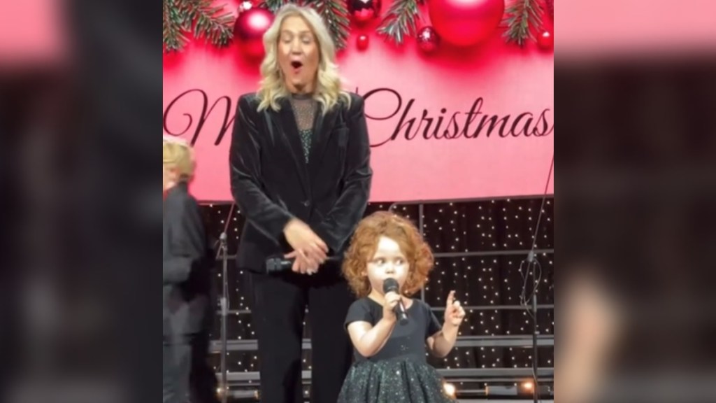A little girl stands on a stage, mic in hand, as she talks. With one hand she holds a mic and with the other, she holds up one finger. A woman stands next to her, mouth wide open and eyes closed, shocked by what is being said. Behind them on stage is a Christmas banner