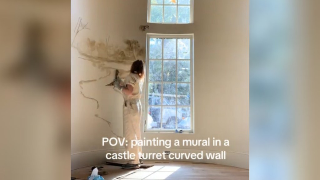 View from a distance of a woman painting a mural on a curved wall. At the center of the wall are two windows