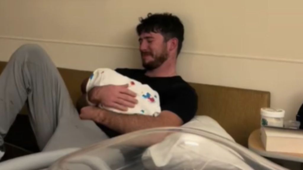A man crying while holding a swaddled baby in his arms.