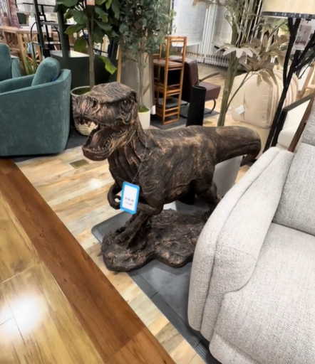 A large dinosaur statue is sat in a HomeGoods next to a neutral chair, a lamp, and a fake plant