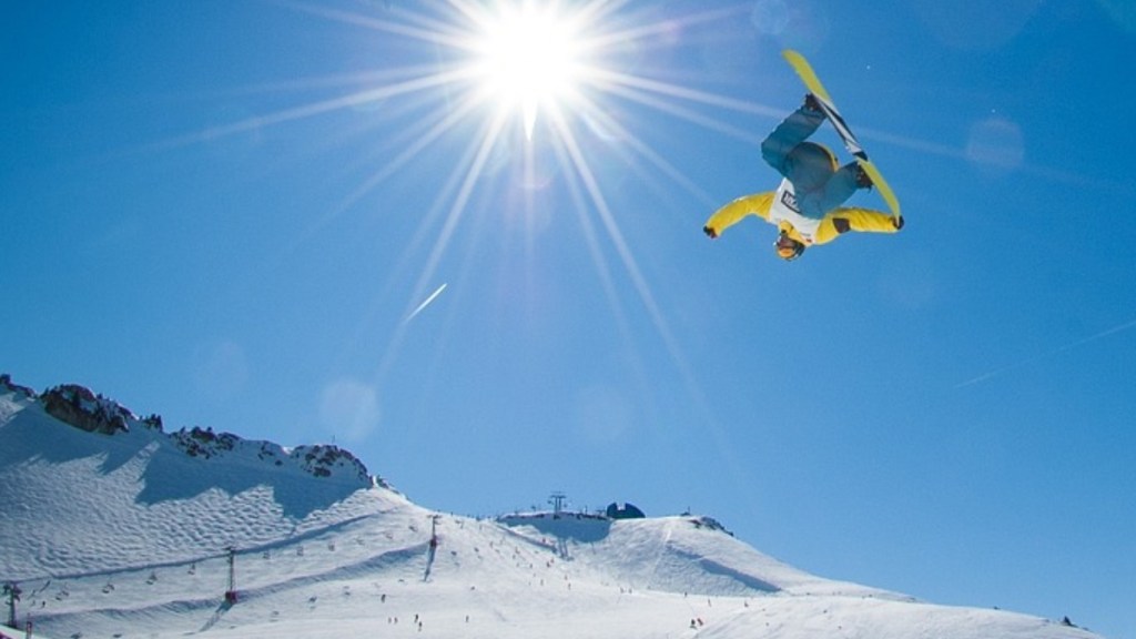 View from afar of a snowboarder soaring through the air, upside down. The sun shines brightly behind him. Snow-covered hills are below.