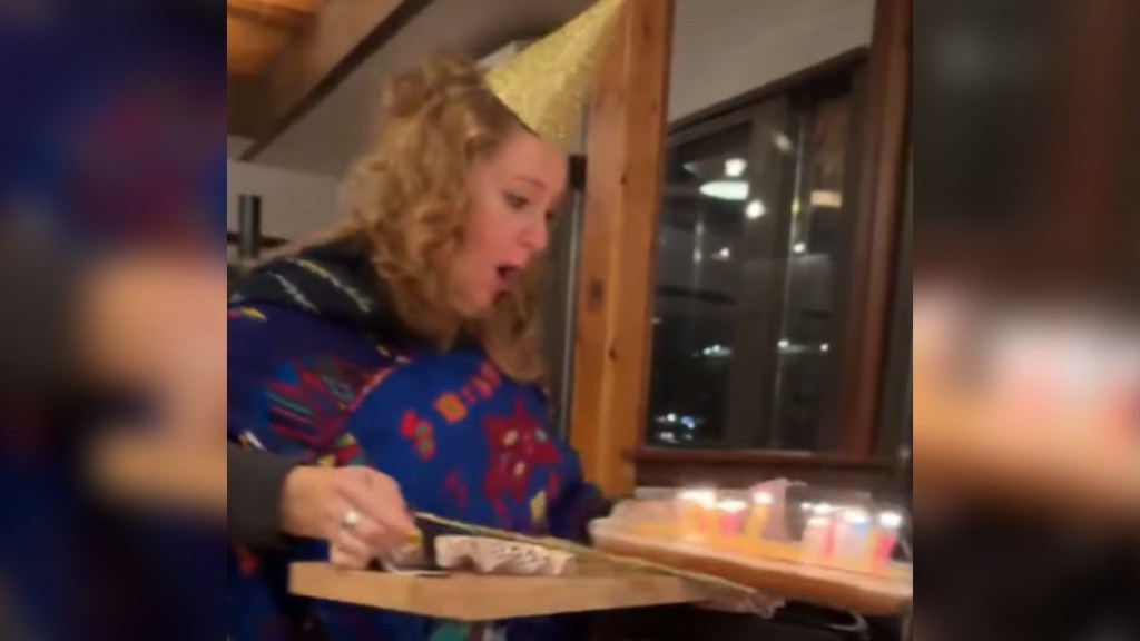 A woman looks down, eyes wide, and mouth open, at the tray she's carrying. On the tray is a birthday cake, with lit candles, that's slipping off
