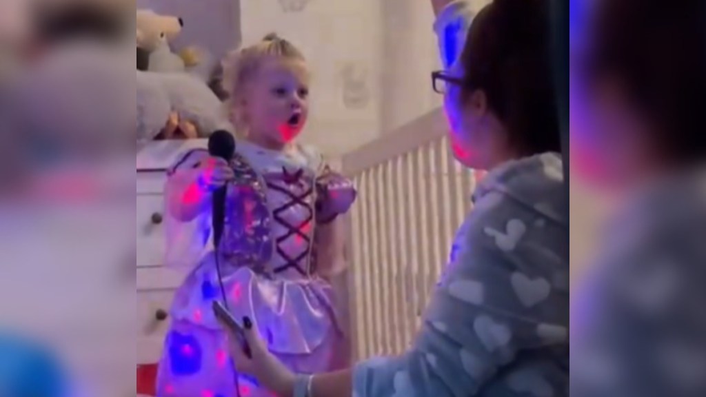 A mom and little girl sit on the floor, facing each other. We only see the side of mom who is raising one hand in the air as she sings. The little girl can be seen with her mouth wide open as she sings