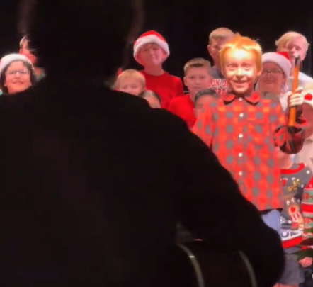 View from behind the shadow of a teacher playing a guitar. He is approaching a stage full of kids. One of the kids smiles wide as he holds up a hammer