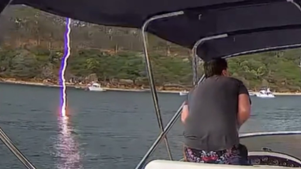 View from behind of a man driving a boat. Feet away, a lightning strike is hitting the water