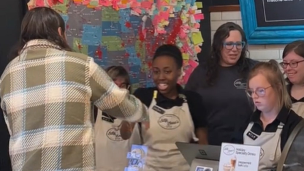 View from behind of a man standing at a coffee shop counter, checking out. One employee smiles as she goes to shake his hand. Four others stand nearby, happily watching