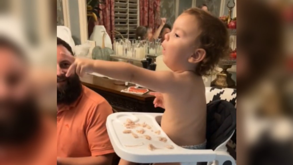 A baby sits in his high chair at a dining table. He enthusiastically points to something in the distance as a man sitting next to him smiles at him