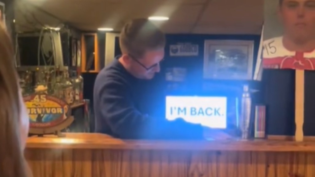 A young man stands behind a counter, looking down at a laptop with a PowerPoint presentation open. The screen reads: I'M BACK