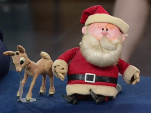 Original Rudolph and Santa puppets are sat on a table with a blue tablecloth. They're both damaged from a fire
