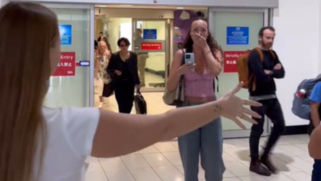 View from behind a woman who has an arm stretched out wide to hug the woman walking toward her. This woman is crying. She covers her mouth with one of her hands.