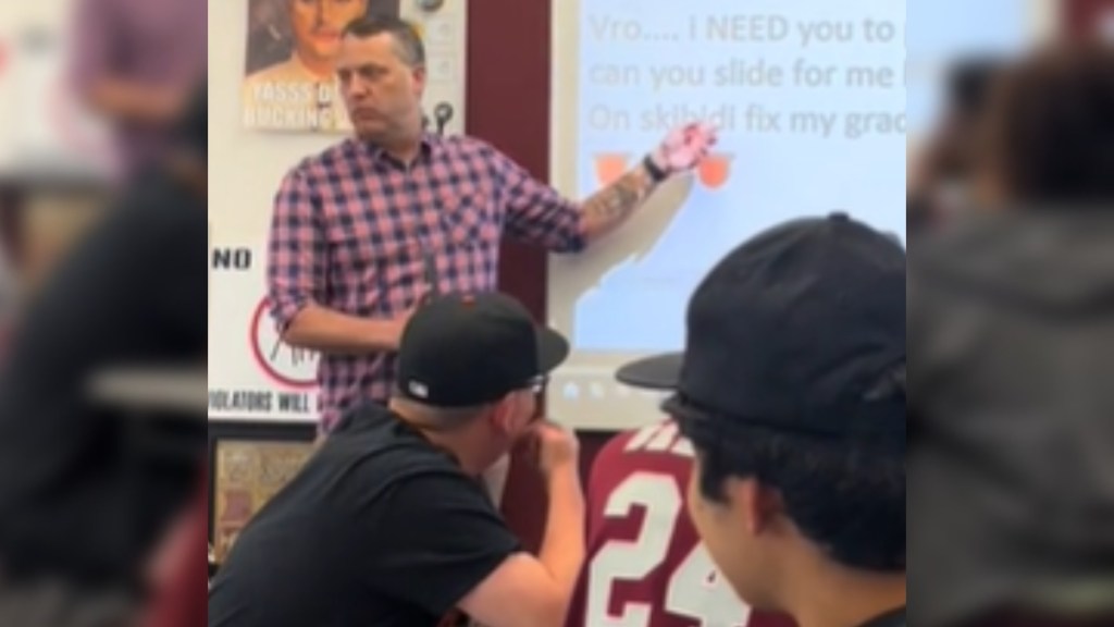 From the perspective of a student, a teacher stands at the front of a classroom. He talks as he points to something being projected onto a screen
