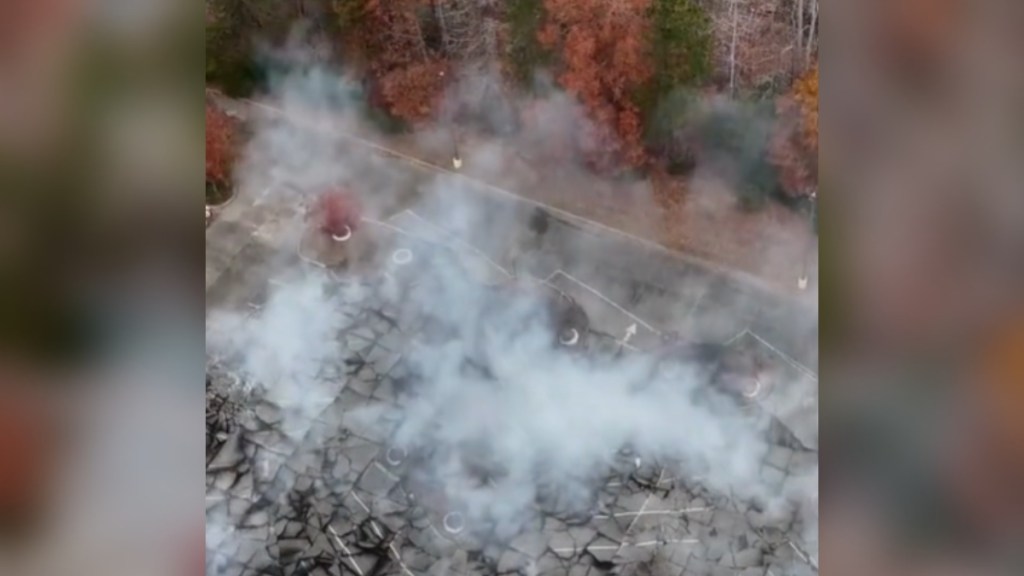 Aerial view of a parking lot with a fire beneath. Smoke rises from the parking lot. Nearby are colorful fall trees