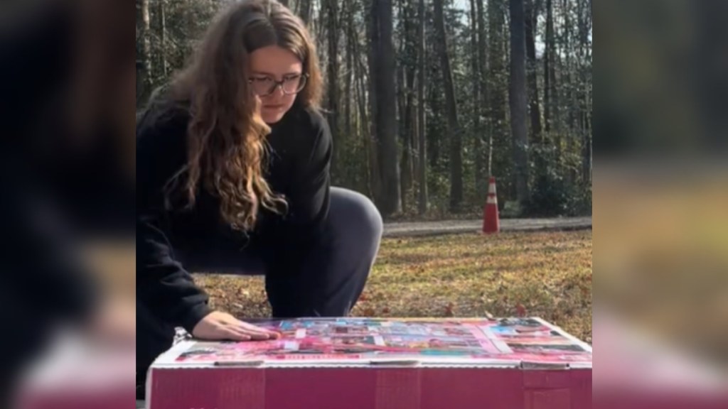 A woman squats near a giant Barbie Dream House box outside.