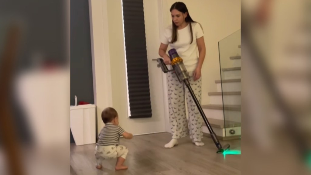 A mom stands with a vacuum in hand. She looks down in shock and awe at her baby who is starting to stand
