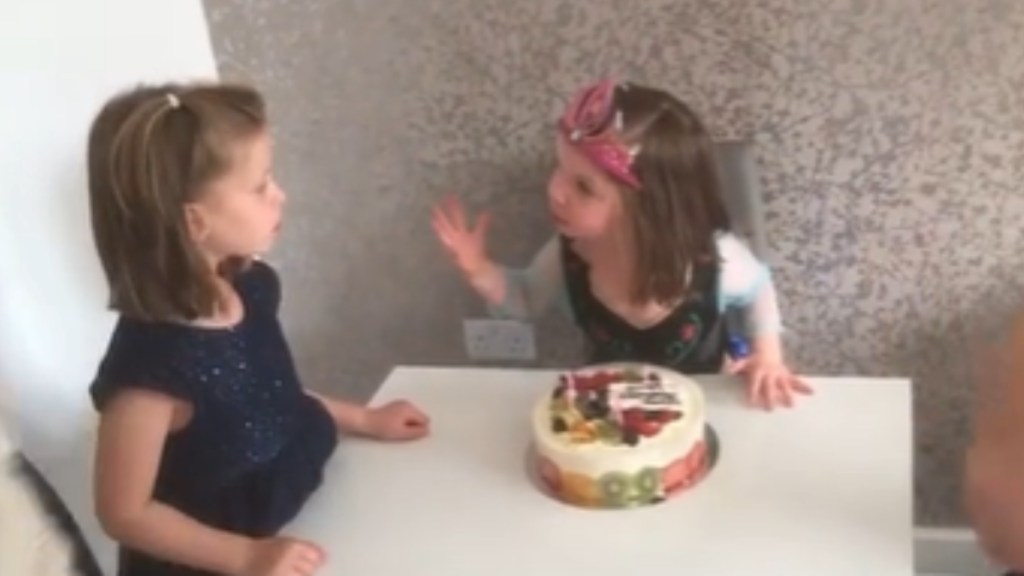 Two little girls sit at a small dining table. One wears a crown and uses her hands as she talks intensely to the other little girl. The other girl stands still, staring blankly at the girl in the crown. A cake sits on the table, no candles lit