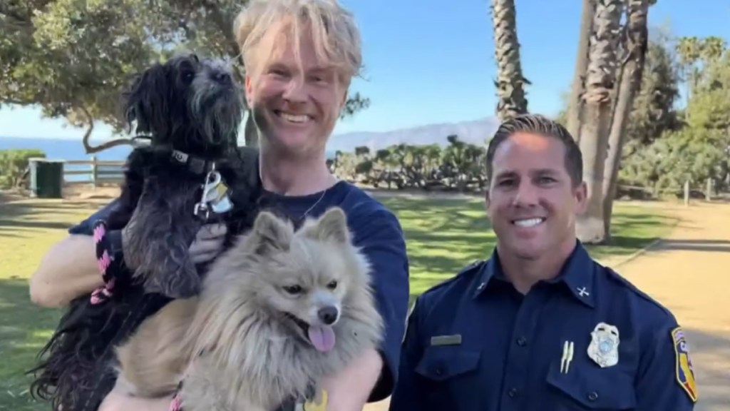Casey Colvin and firefighter Brent Pascua smile wide as they pose for a photo outside. Casey holds both of his dogs, Tika and Oreo, in his arms.