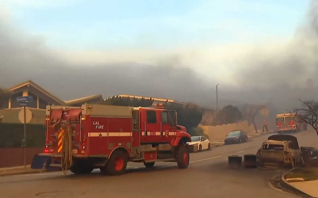 View of a neighborhood street full of smoke from nearby wildfires in Los Angeles. Two firetrucks are parked on the side of the road