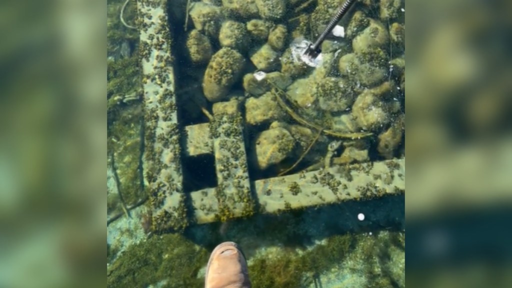 View from someone looking down at a frozen lake. Part of the camera person's shoe can be seen. The clear, frozen water shows lots of rock below the surface. A stick is firmly on the ice, slightly breaking the frozen surface