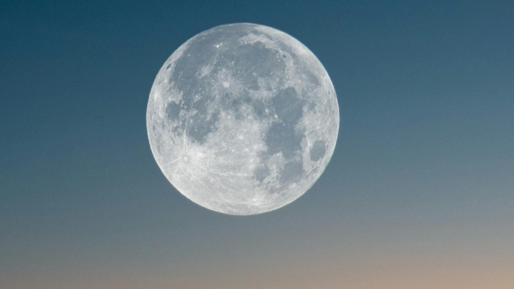 Close up of a full moon. The sky is a gradient of medium to light blues and light orange