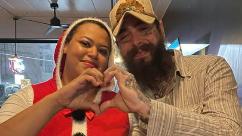 Close up of a woman smiling and posing with Post Malone. With one hand each, they make the shape of a half a heart. Together, their hands make one heart.