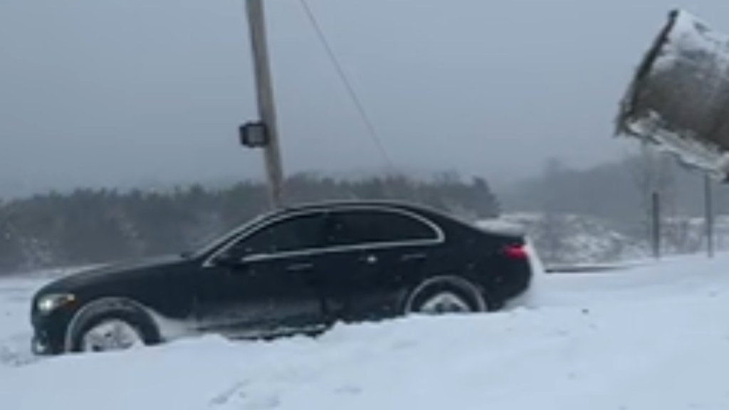 A black car is stuck in snow. A nearby object, a large bale of hay, is being lifted in the air by something that can't be seen in this image just behind it