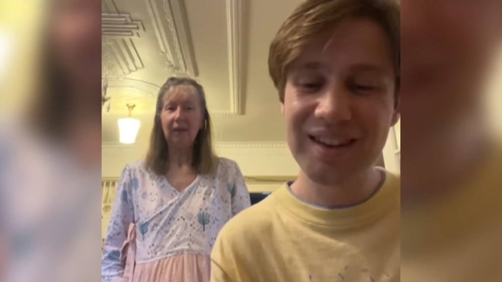 Close to the camera, a man smiles as he sings. Standing a bit behind him, an older woman sings along, looking at the camera