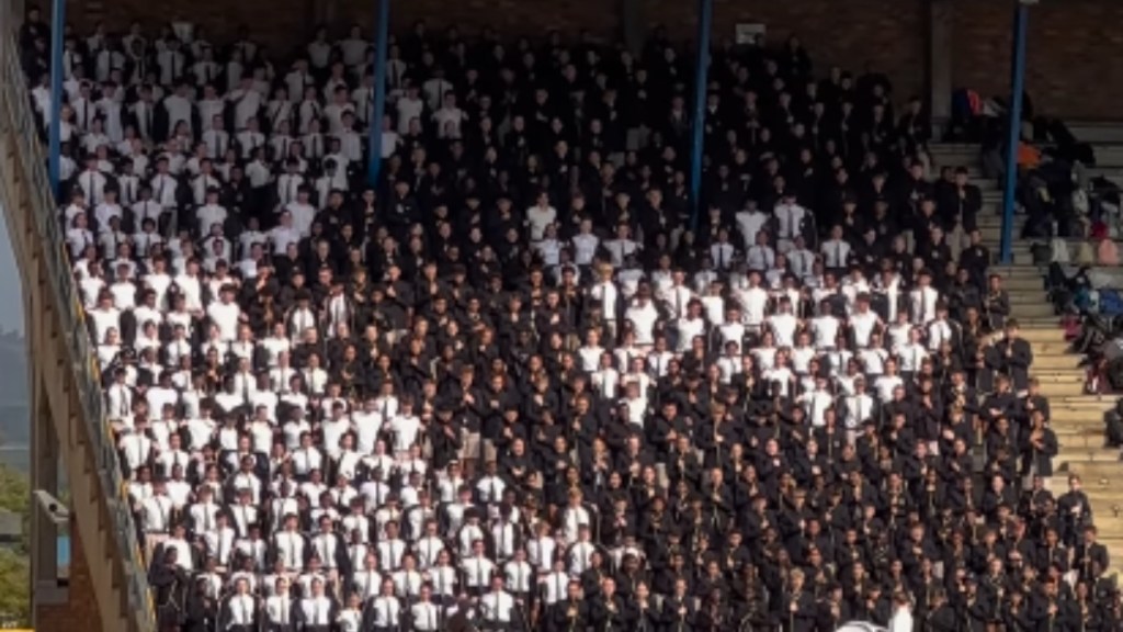 Close up of a large group of high school performers standing outdoors in a stadium. Some are dressed in black while others are in white. Those in white are standing to where an image is being made. The image is of a small fish running away from a larger fish with its mouth wide open
