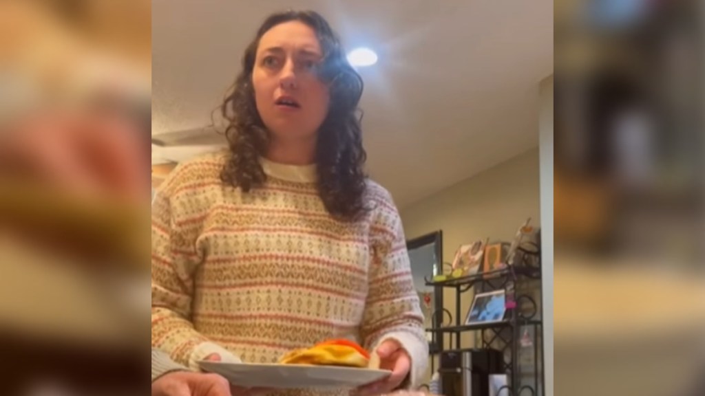 A young woman stands in a kitchen, plate with food in her hand. She looks up at someone who can't be seen in this image. She looks shocked and upset, eyes wide and mouth agape.