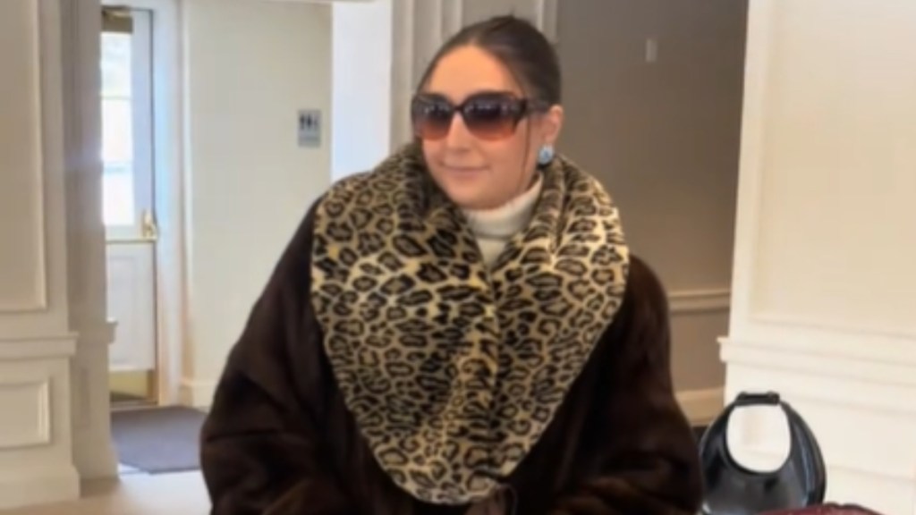Close up of a woman dressed up in a coat with a large leopard-patterned collar. She has her hair pulled back and wears large sunglasses. She smiles coyly as she poses