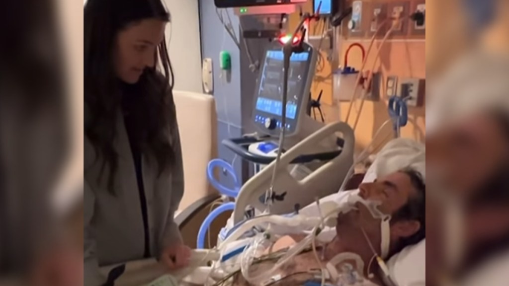 A man in a coma lays in a hospital bed, hooked up to machines. A woman stand next to the bed, looking down at him
