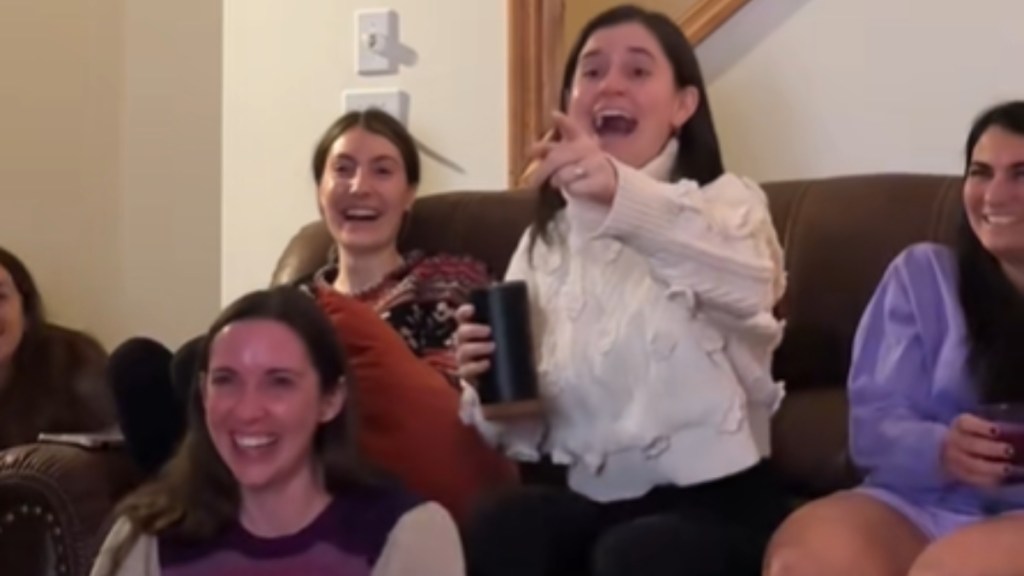 A group of five women sit on a couch and the floor. One woman in the middle sits on the edge of the couch, happily shocked. She points at something that can't be seen in this image, eyes wide, mouth wide open. The women around her look just as excited