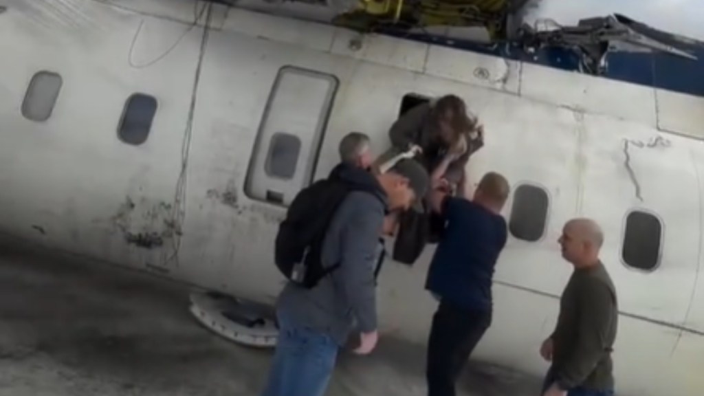 A plane has landed upside down. Three people stand outside, assisting a woman as she exits the plane