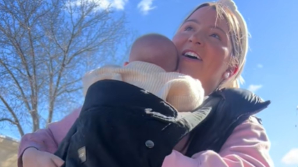 Close up of a smiling woman who holds a baby in a carrier on her chest