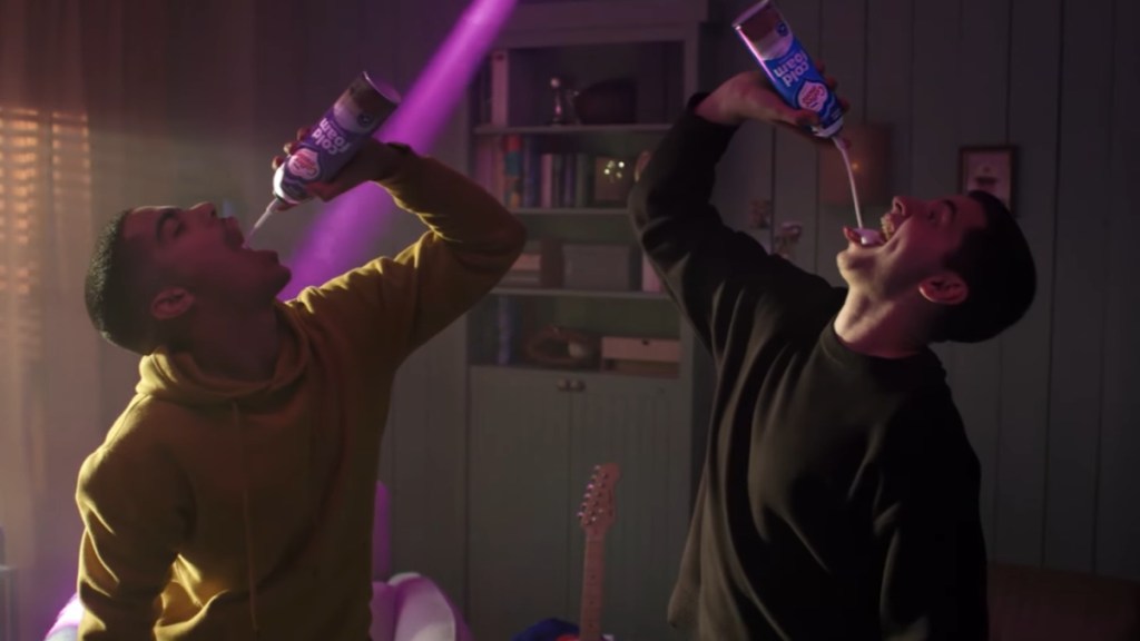 Two men stand next to each other, mainly facing the camera. Each holds a Coffee Mate Cold Cream Foam in the air, mouths open as they eat some. A bright purple light shines in the background for dramatic effect