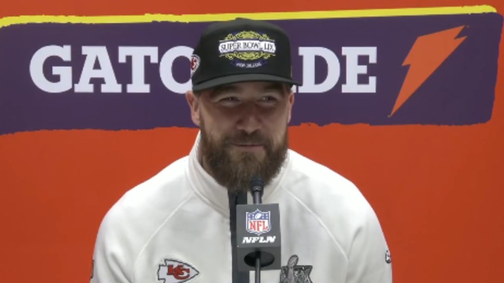 Travis Kelce sits in front of a mic for media interviews. He smiles