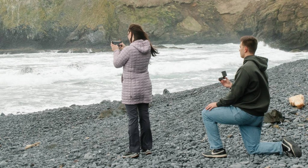 A man and woman are on a beach near the water. The man waits on on knee, engagement ring out. The woman has her back to him. Her phone is out and she is focused on taking a picture. She is facing the water.