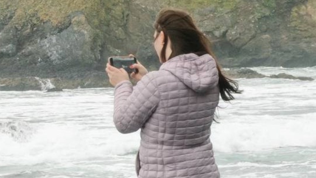 Close up of a woman standing on the shore. Her back is mainly toward the camera. She has her phone out and is taking a photo of something closer to the water