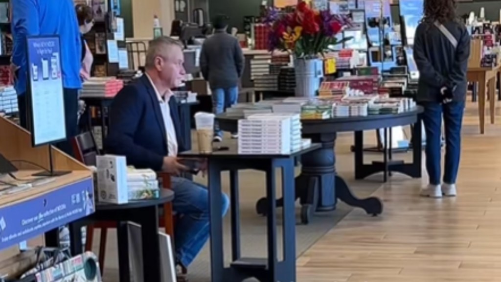 Inside a Barnes & Noble, a man sits at a table with stacks of books in front of him. He looks around, phone in one hand at his lap. People are kind of nearby, but none of them are at his table